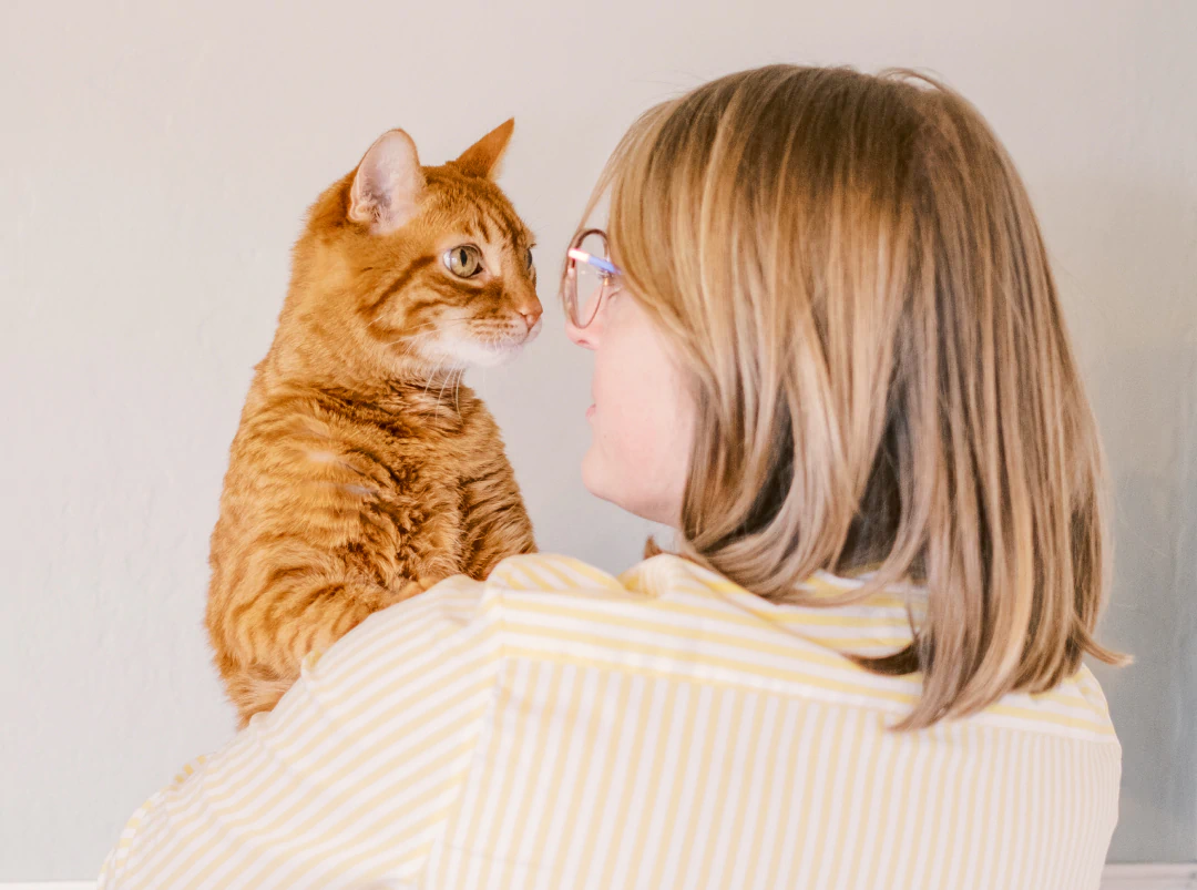 Desktop_About_Us_How_We_Approach A person with shoulder-length blonde hair and glasses, wearing a yellow and white striped shirt, holds an orange tabby cat while they look at each other.