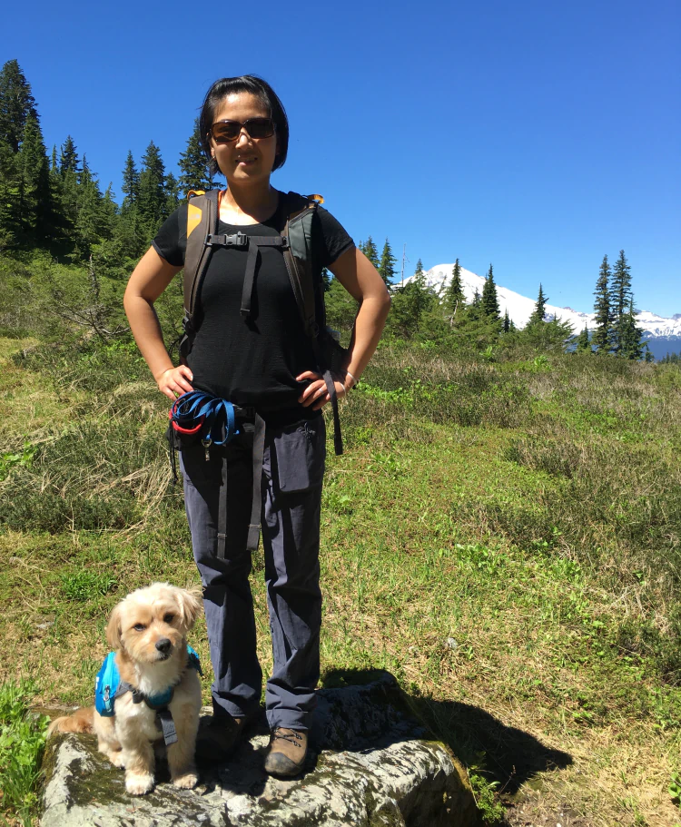 Desktop_Meet_the_Doctor_Her_Philosophy_1 A woman wearing sunglasses and hiking gear stands on a grassy field with a small dog in a blue vest sitting on a rock in front of her. There are trees and snow-capped mountains in the background under a clear blue sky.