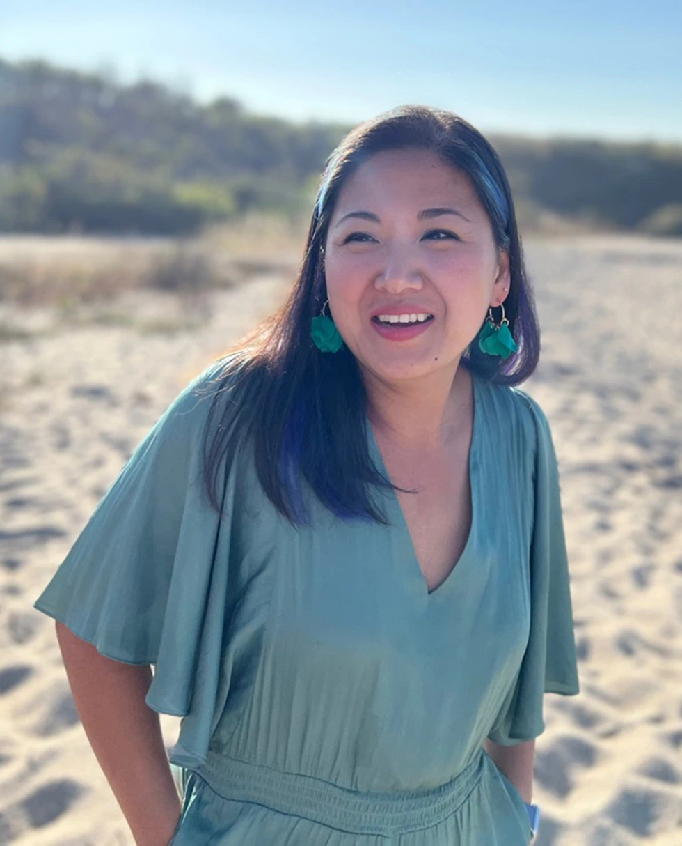 Desktop_Meet_the_Doctor_Meet_Dr_Guan A woman with long dark hair and green earrings smiles while standing on a sandy beach, wearing a light green dress. The background features blurred trees and a bright, clear sky.